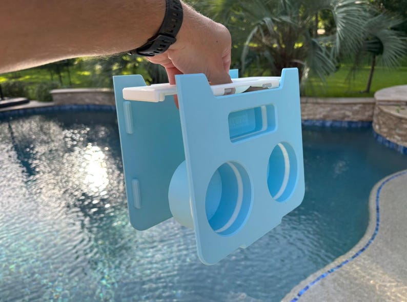 Op de afbeelding: Een lichtblauwe plastic pool float houder met twee ronde bekerhouders en een centrale cilindrische sectie. De houder wordt door een hand omhoog gehouden. De achtergrond is een zwembad.