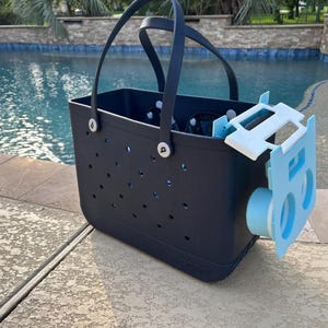 Op de afbeelding: Een marineblauwe draagtas met een lichtblauw accessoire bevestigd. De tas heeft een geperforeerd ontwerp en zwarte handvatten. Het accessoire is lichtblauw en wit, met een cilindrisch onderdeel. De achtergrond toont een zwembad.