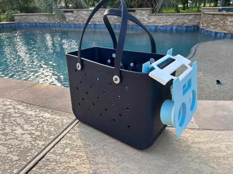 Op de afbeelding: Een marineblauwe draagtas met een lichtblauw accessoire bevestigd. De tas heeft een geperforeerd ontwerp en zwarte handvatten. Het accessoire is lichtblauw en wit, met een cilindrisch onderdeel. De achtergrond toont een zwembad.