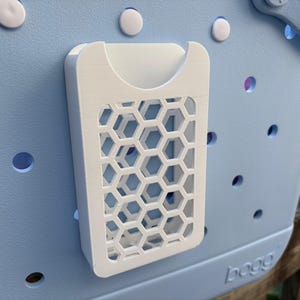 May include: A white, 3D-printed organizer with a honeycomb pattern attached to a light blue tote bag. The organizer has a pocket and a geometric design. The bag has several round holes and white fasteners.