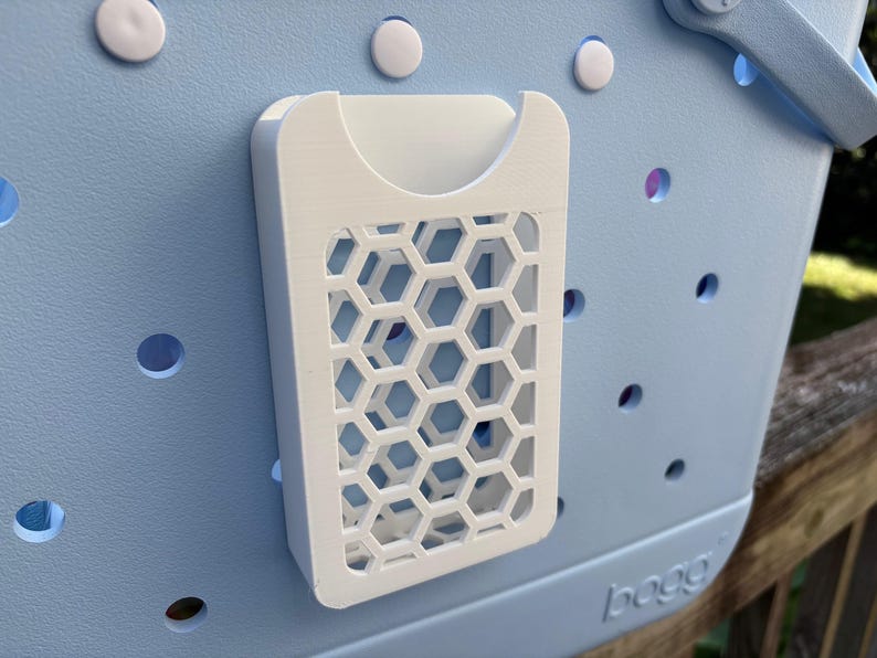 May include: A white, 3D-printed organizer with a honeycomb pattern attached to a light blue tote bag. The organizer has a pocket and a geometric design. The bag has several round holes and white fasteners.
