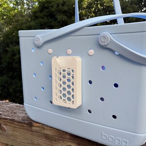 May include: A light blue tote bag with a white honeycomb-patterned accessory attached. The bag has a handle and small holes throughout. The word "bogg" is printed on the bottom of the bag.