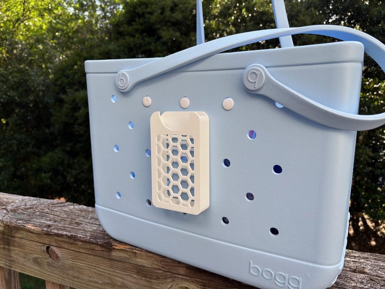 May include: A light blue tote bag with a white honeycomb-patterned accessory attached. The bag has a handle and small holes throughout. The word "bogg" is printed on the bottom of the bag.