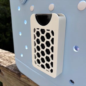May include: A light blue tote bag with a white, honeycomb-patterned accessory attached. The accessory has a black interior and is mounted on the bag's exterior. The bag features several round, white accents and circular cutouts.