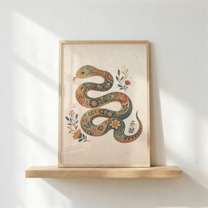 May include: A framed rectangular wall art print featuring a winding snake with a detailed floral and paisley pattern in shades of orange, teal, and cream. The snake is surrounded by small botanical sprigs and flowers on a light beige background.