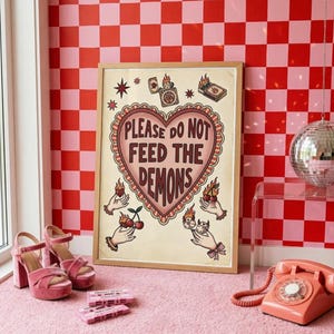 Könnte beinhalten: Ein gerahmter Druck mit dem Text "PLEASE DO NOT FEED THE DEMONS" in Herzform, mit Illustrationen von Händen, die Kirschen und Flammen halten. Das Bild ist vor einer rot-rosa karierten Wand platziert, mit rosa Samt-Plateau-Absätzen und einem rosa Wählscheibentelefon.