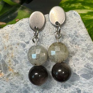 May include: A pair of silver earrings with two round beads. The top bead is a light grey colour with a translucent, speckled appearance. The bottom bead is a dark grey colour with a smooth, shiny finish.