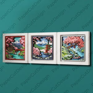 May include: Three framed paper cut art pieces showing Japanese landscapes. Each piece features layered designs with cherry blossoms, mountains, pagodas, and water features. The frames are white, and the background is teal.