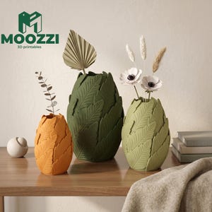 Può includere: Tre vasi decorativi arancioni, verdi e verde oliva, ciascuno con un design a forma di foglia. I vasi sono su una superficie di legno e contengono fiori e foglie secche. Il logo "MOOZZI 3D printables" è visibile.