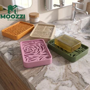 Puede incluir: Cuatro jaboneras rectangulares en naranja, beige, rosa y verde oliva, cada una con un patrón de corte geométrico o floral único. Una pastilla de jabón amarilla translúcida descansa sobre la jabonera verde oliva. El logo MOOZZI 3D printables es visible.