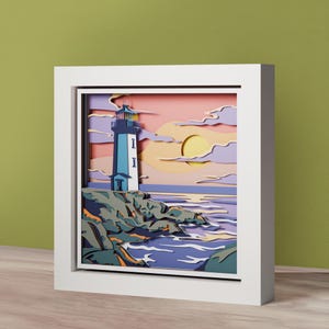 Puede incluir: Una obra de arte en papel en capas que representa un faro en una costa rocosa al atardecer. El faro es blanco y azul, sobre un cielo rosa y amarillo con un gran sol. La obra de arte está enmarcada en una caja de sombra blanca.