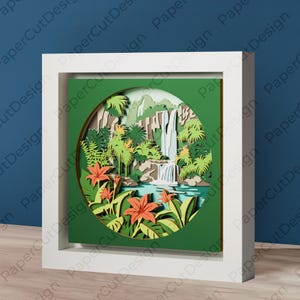 Może przedstawiać: Oprawiony projekt wycinanki z papieru przedstawiający warstwową scenę tropikalnego wodospadu. Grafika zawiera wodospad, bujne zielone liście i pomarańczowe kwiaty, a wszystko to w okrągłej ramie. Rama jest biała, a tło jest głęboko zielone.