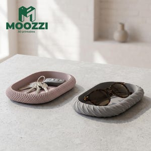 Puede incluir: Dos bandejas organizadoras ovaladas. Una bandeja es de color rosa empolvado con un borde texturizado de patrón de diamantes y contiene un juego de llaves. La segunda es de color gris claro con un borde de textura estriada y contiene gafas de sol.