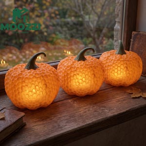 Puede incluir: Tres calabazas naranjas con una superficie texturizada de panal y tallos de color verde oscuro. Cada calabaza está iluminada desde el interior, emitiendo un brillo cálido. Las calabazas están dispuestas en fila sobre una superficie de madera.