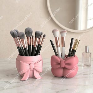 Puede incluir: Dos recipientes rosas en forma de lazo que contienen brochas y aplicadores de maquillaje. Los recipientes están sobre una superficie de mármol, con un espejo y un frasco de perfume en el fondo. Las brochas tienen mangos negros y detalles en oro rosa.