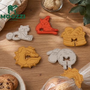 Op de afbeelding: Een verzameling kleurrijke, diervormige zakclips, waaronder een kat, hond, schaap en broodmand. De clips zijn gemaakt van plastic en zijn ontworpen om zakken af ​​te sluiten. Het logo "MOOZZI 3D printables" is zichtbaar.