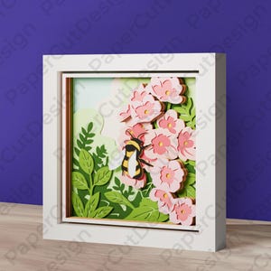 May include: A framed paper cut artwork depicting a bee on pink flowers. The layered design creates a 3D effect. The bee has black and yellow stripes. Green leaves and a light blue background complete the scene.