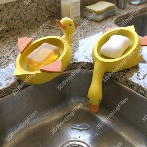 May include: Two yellow duck-shaped soap dishes with orange beaks and feet. One holds a clear bar of soap, the other a white bar. The dishes are placed near a stainless steel sink.