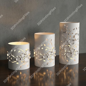 May include: Three white cylindrical candle holders with floral cutouts. The candle holders are of varying heights and feature intricate designs of flowers and leaves. The cutouts allow light to shine through, creating a warm glow. The candle holders are on a dark wooden surface.