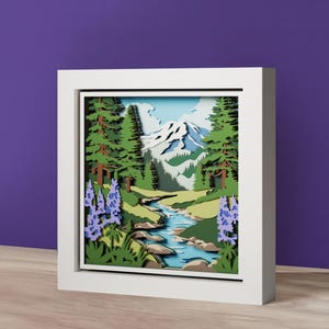 Può includere: Un'opera d'arte in carta incorniciata raffigurante un paesaggio montano a strati. L'opera presenta un fiume, alberi e montagne nei toni del verde, blu e bianco. La cornice bianca esalta l'effetto 3D.