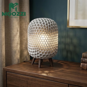 May include: A table lamp with a cylindrical, white, honeycomb-patterned shade that emits a warm, glowing light. The lamp is supported by three short, dark brown wooden legs and includes a black power cord extending from the base.