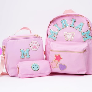 May include: A pink backpack, lunch bag, and pencil case set. The backpack has the name "MIRIAM" in blue sequined letters. The lunch bag has a paw print and the letter "M". The pencil case has a smiley face.
