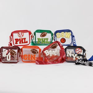 May include: A collection of clear, rectangular crossbody bags with various team names and sports-themed designs. Each bag features a different color scheme, including red, green, and blue, with a baseball or football graphic. Some bags have matching straps with text like "GAME DAY".