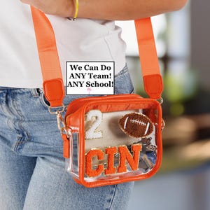 May include: Clear orange-trimmed crossbody bag with a matching strap. The bag features a football-shaped patch, the number 2, and the letters CIN. The text "We Can Do ANY Team! ANY School!" is visible.