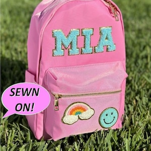 May include: Pink backpack with a blue and gold "MIA" name patch, a rainbow and cloud patch, and a smiley face patch. The text "SEWN ON!" is in a speech bubble.