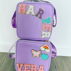 May include: Two lavender lunch bags with shoulder straps. One bag has the name "HARPER" in colorful, glittery letters. The other bag has the name "VERA" and patches of a butterfly, rainbow, and heart.