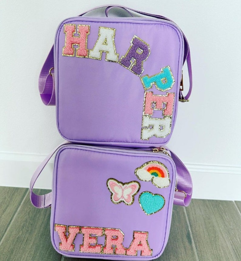 May include: Two lavender lunch bags with shoulder straps. One bag has the name "HARPER" in colorful, glittery letters. The other bag has the name "VERA" and patches of a butterfly, rainbow, and heart.