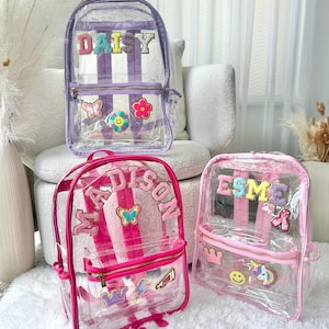 Puede incluir: Tres mochilas transparentes con nombres personalizados y parches decorativos. Las mochilas presentan rayas verticales y detalles coloridos. Una mochila tiene el nombre "DAISY" y rayas moradas, otra "ADDISON" con rayas rosas, y la tercera "ESME" con rayas rosas.