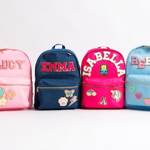 May include: Four colorful backpacks with personalized names. Pink backpack with "LUCY" and flower patches. A blue denim backpack with "EMMA" and patches. A hot pink backpack with "ISABELLA" and patches. A light blue backpack with "REESE" and patches.