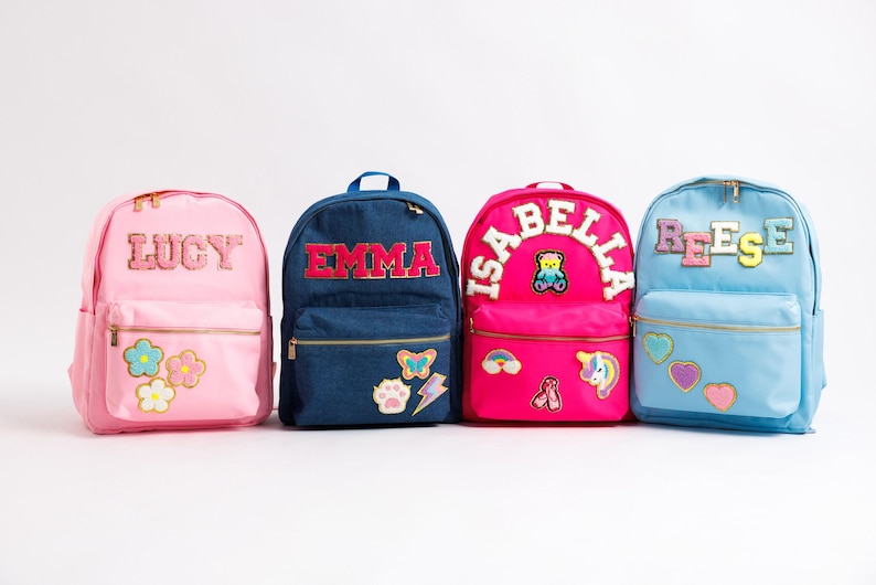 May include: Four colorful backpacks with personalized names. Pink backpack with "LUCY" and flower patches. A blue denim backpack with "EMMA" and patches. A hot pink backpack with "ISABELLA" and patches. A light blue backpack with "REESE" and patches.