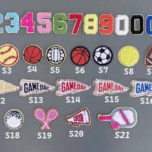 May include: A collection of embroidered patches featuring numbers, sports balls, and pennants. The patches include numbers 0-9, soccer balls, baseballs, basketballs, tennis balls, footballs, and pennants with "GAME DAY" text.