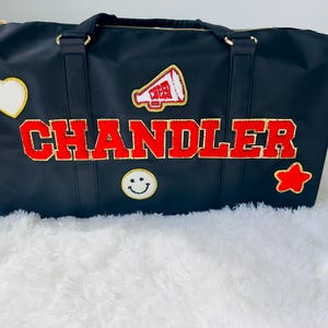 May include: Black duffel bag with gold hardware and the name "CHANDLER" in red varsity letters. The bag features a heart, star, and smiley face patch. A megaphone patch with the word "CHEER" is also visible.