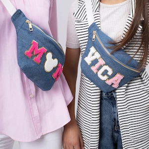 May include: Two denim fanny packs with white straps. One fanny pack has pink and white lettering that says "Mickey" and the other has pink and white lettering that says "Vica".
