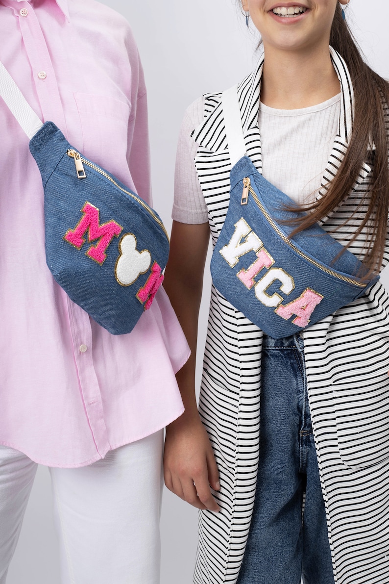 May include: Two denim fanny packs with white straps. One fanny pack has pink and white lettering that says "Mickey" and the other has pink and white lettering that says "Vica".