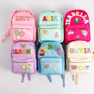 May include: Six colorful backpacks with personalized names, including "LUCY," "ARIA," and "ISABELLA." Each backpack features a front pocket, adjustable straps, and decorative patches like flowers, rainbows, and butterflies. The backpacks are in shades of pink, mint green, purple, blue, and cream.