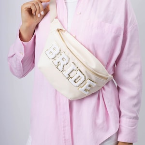 May include: A white fanny pack with a gold zipper and the word "BRIDE" in white letters with gold trim. The fanny pack is worn by a person wearing a pink button-down shirt and white pants.