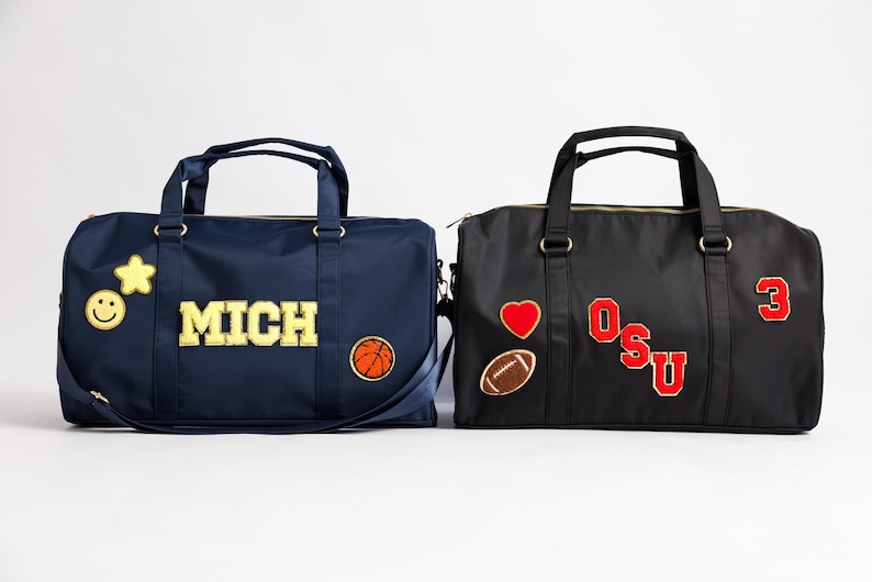 以下が含まれることがあります： 2つのダッフルバッグ。1つは「MICH」の文字、バスケットボール、星、スマイリーフェイスが付いたネイビーブルー。もう1つは「OSU」の文字、ハート、フットボール、数字の3が付いた黒色です。