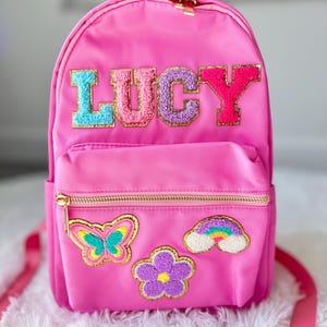May include: A pink backpack with a gold zipper and a fluffy, colorful patch that says "LUCY". The backpack also has a butterfly, flower, and rainbow patch.