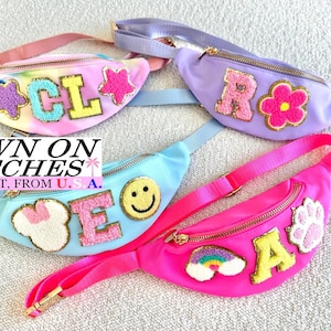 May include: Four colorful fanny packs with embroidered patches. The fanny packs are in pink, light blue, and purple. Each pack has a different design, including letters, stars, a flower, a paw print, a rainbow, and a smiley face. The text "SEWN ON PATCHES" is visible.