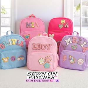 May include: Five colorful children's backpacks in shades of pink, blue, and purple. Each backpack features a name patch and decorative patches like butterflies, flowers, and a unicorn. The backpacks are displayed on a white rug.