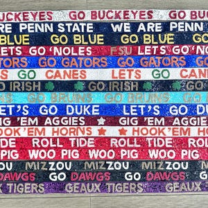May include: A collection of 16 colorful, glittery straps with various college team slogans, including "Go Buckeyes", "We Are Penn State", "Go Blue", "FSU Lets Go 'Noles", "Go Gators", "Lets Go Canes", "Go Irish", "Let's Go Duke", "Gig 'Em Aggies", "Hook 'Em Horns", "Roll Tide", "Woo Pig", "Mizzou", "Go Dawgs", and "Geaux Tigers".