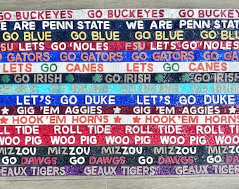 May include: A collection of 16 colorful, glittery straps with various college team slogans, including "Go Buckeyes", "We Are Penn State", "Go Blue", "FSU Lets Go 'Noles", "Go Gators", "Lets Go Canes", "Go Irish", "Let's Go Duke", "Gig 'Em Aggies", "Hook 'Em Horns", "Roll Tide", "Woo Pig", "Mizzou", "Go Dawgs", and "Geaux Tigers".