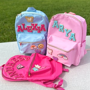 May include: Three colorful backpacks in light blue, pink, and hot pink. Each backpack features a name in a varsity-style font, with decorative patches including a butterfly, rainbow, and smiley face. The bags have gold zippers and are designed for children.
