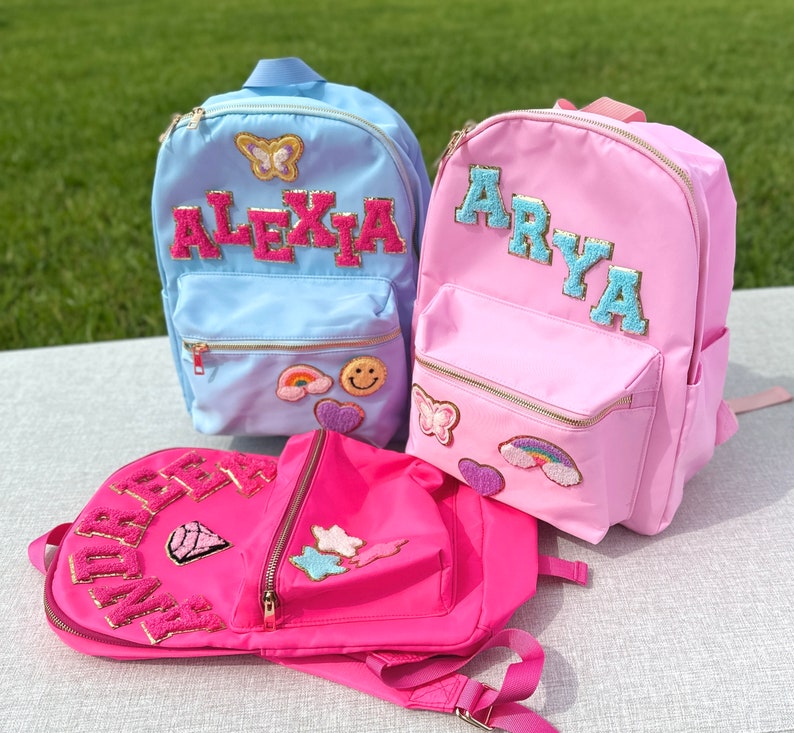 May include: Three colorful backpacks in light blue, pink, and hot pink. Each backpack features a name in a varsity-style font, with decorative patches including a butterfly, rainbow, and smiley face. The bags have gold zippers and are designed for children.