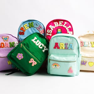 May include: A collection of colorful backpacks, each with a name embroidered on the front. The backpacks are in various colors, including pink, green, blue, and light blue. Decorative patches, such as flowers, rainbows, and crowns, adorn the bags.
