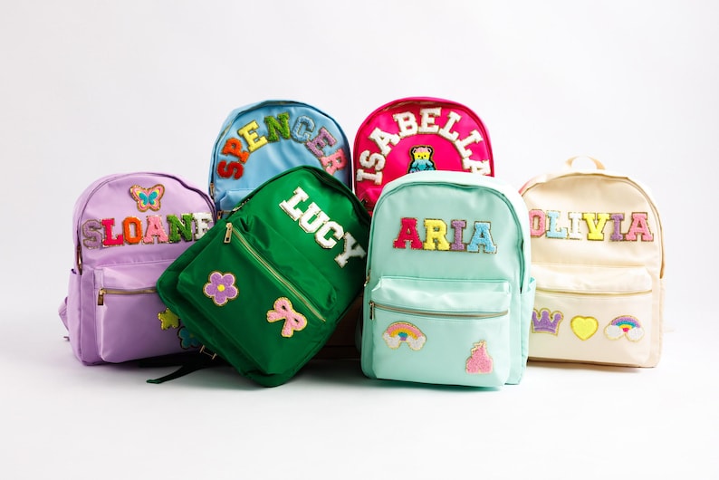 May include: A collection of colorful backpacks, each with a name embroidered on the front. The backpacks are in various colors, including pink, green, blue, and light blue. Decorative patches, such as flowers, rainbows, and crowns, adorn the bags.